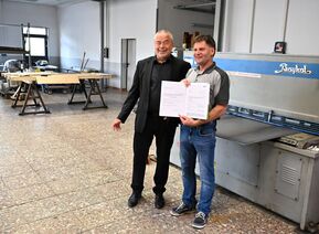 zwei Männer stehen vor einer Maschine und halten einen Bescheid in der Hand 