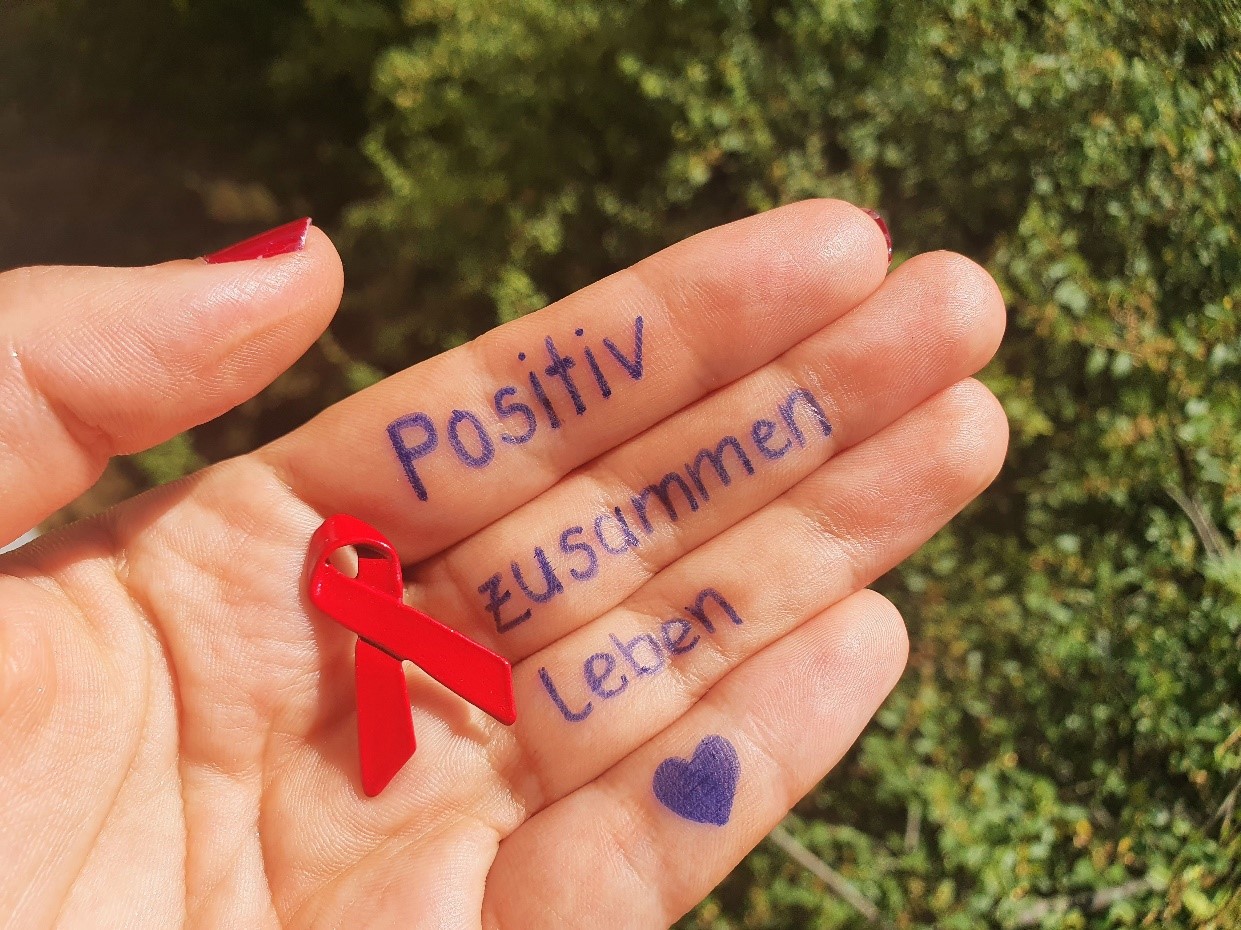 Hand mit roter Schleife und Schrift "positiv zusammen leben" Herzchen auf dem kleinen Finger