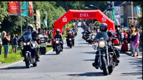 Motorradfahrer und -fahrerinnen auf abgesperrter Straße mit Personen am Rand 